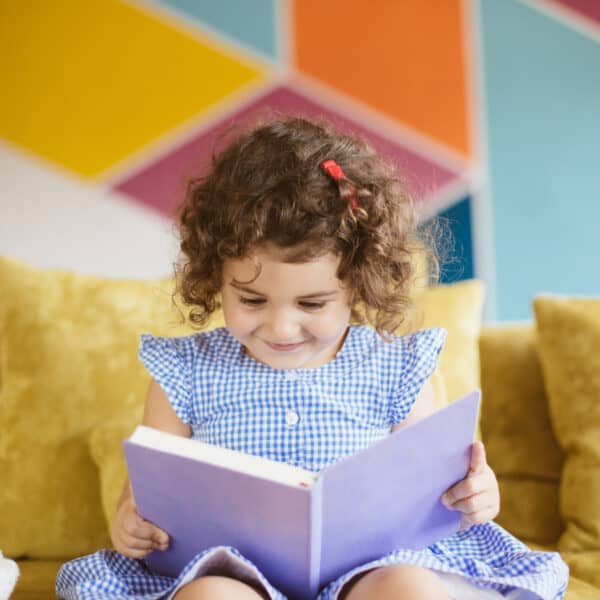 Beautiful little smiling girl with dark curly hair in blue dress reading child book on sofa at home